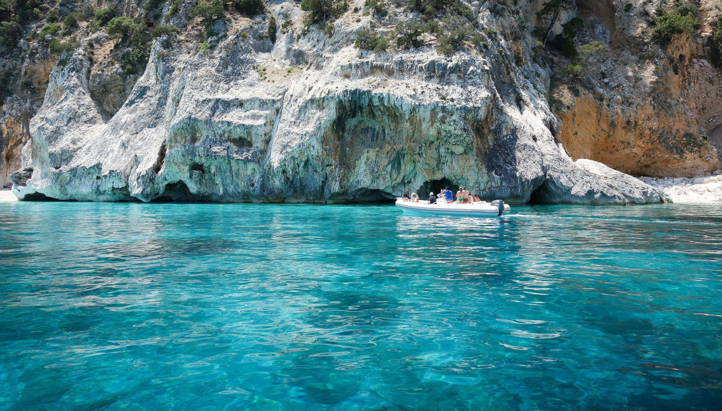 A small private boat with guests in a turquoise cove beneath pale limestone cliffs